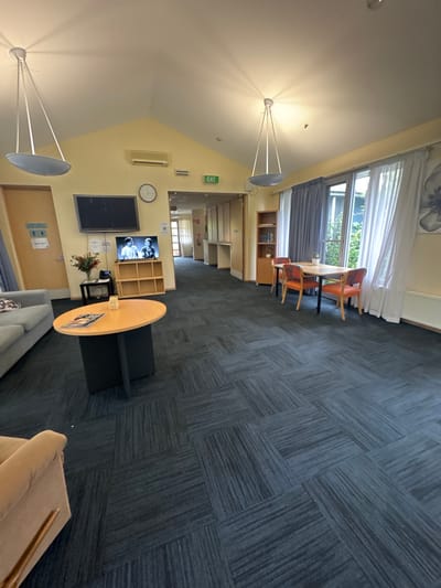 This image captures a well-lit, unoccupied indoor common area or lounge, possibly within a community center or care facility in Carrum Downs, Australia. The floor is covered with dark grey carpet tiles laid in a checkerboard pattern, featuring alternating stripe directions.

Along one side of the room, large windows are adorned with white sheer curtains and grey blackout curtains, revealing green foliage outside. In front of these windows, a wooden dining table is surrounded by four chairs upholstered in orange. Nearby, a wooden built-in cabinet or shelving unit is visible.

The central part of the room functions as a lounge area, with a light-colored sofa and a beige armchair partially seen. A round wooden coffee table with a black base sits in front of these seating arrangements. Mounted on a light yellow wall is a large flat-screen television, currently displaying a black and white film or program featuring two men. Adjacent to the TV is a light wooden shelving unit. A wall clock and an air conditioning unit are positioned above the television.

The room features a high, vaulted white ceiling from which two modern pendant lights are suspended, casting distinct radial light rays. A green "EXIT" sign is prominently displayed on the wall. Further into the background, a corridor leads to additional interior spaces and doorways. The presence of natural light through the windows suggests it is daytime. No people are present, indicating a quiet moment in the facility.