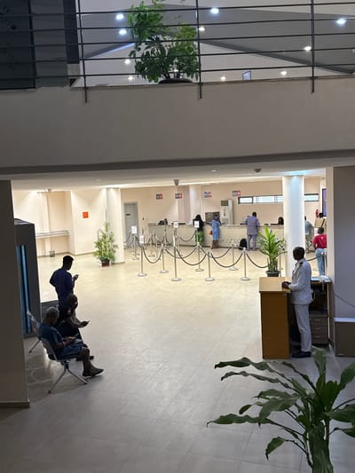 The image depicts an indoor scene, likely a government building or a public service office in Lagos, Nigeria. The setting is a long hallway with a reception or service counter on one side. Several people are present, some standing in line at the counter and others seated in a waiting area.
At the service counter, individuals appear to be interacting with staff, though the faces of the staff are not clearly visible. One person in a white coat is standing at the counter, suggesting they might be an employee. The counter is equipped with stanchions and ropes to manage queues.
In the waiting area, several people are seated, some holding mobile phones and seemingly engaged with them. The overall atmosphere appears to be one of routine activity, with no overt signs of distress or excitement. The lighting suggests it is daytime, possibly late afternoon or early evening, as the interior lights are on.
Plants are visible in pots, adding a touch of greenery to the otherwise functional environment. The architecture features modern elements with tiled floors and smooth walls. The image captures a slice of everyday life in a public service setting.