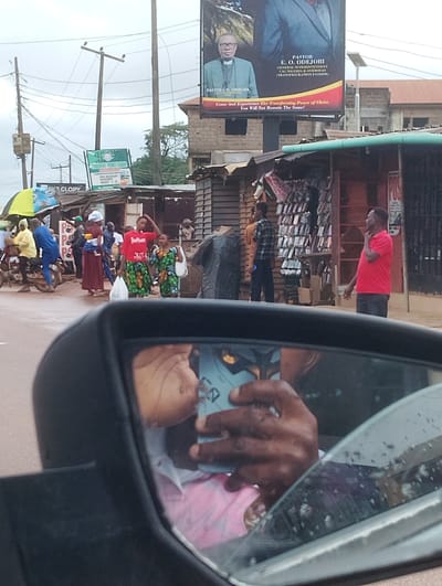 This is a medium-shot, eye-level photograph taken from inside a vehicle, looking out of a side mirror and through the windshield. The image captures a street scene in Aketu-Oja, Nigeria.
In the foreground, a black side mirror is prominent, reflecting a person's hands holding a smartphone and a glimpse of their face. The background reveals a busy street with several people, stalls, and a large billboard.
The billboard at the top features a portrait of a man identified as "PASTOR E. O. ODEJOBI, GENERAL SUPERINTENDENT, CAC NIGERIA & OVERSEAS (TRANSFIGURATION FATHER)". Below his image, text reads, "Come And Experience The Transforming Power of Christ. You Will Not Remain The Same!".
On the street level, various individuals are present, some gathered around market stalls selling goods. One stall displays an array of phone cases. People are dressed in casual clothing, with some wearing brightly colored fabrics. A woman in the middle ground wears a red t-shirt with "jubilee" printed on it and a green and black patterned skirt. Another woman beside her wears a similar patterned skirt and a white top.
Several utility poles and overhead wires are visible, suggesting an urban or semi-urban environment. The lighting suggests it might be daytime, possibly overcast due to the muted colors and diffused light. The overall scene depicts a lively street market atmosphere in Nigeria.