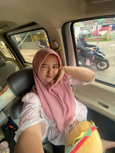 A person wearing a pink hijab and a floral-patterned dress poses inside a car, winking at the camera. A plush burger toy is visible next to them. Outside the car window, a street scene includes a motorcycle with two people nearby, one lying on the ground and another sitting on the bike. The car interior shows gray seats and a window.