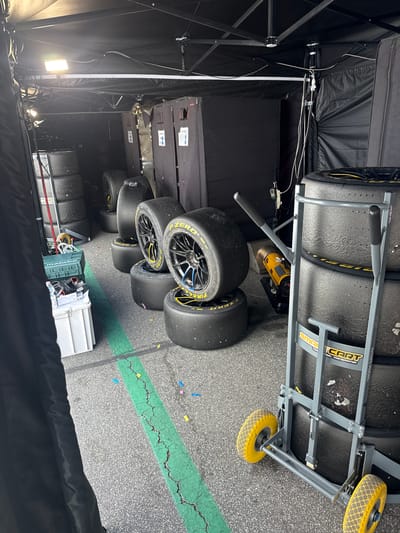 The image shows a Formula 1 pit lane garage, likely located at the Autodromo Vallelunga Piero Taruffi in Campagnano di Roma, Italy, given the presence of Formula 1 tires.  Several stacks of Pirelli P Zero Formula 1 tires are visible, some on a cart. The tires feature yellow sidewall markings.  The garage area has overhead lighting and a black fabric covering some portions of the structure.  Colored confetti is scattered on the asphalt surface of the pit lane.