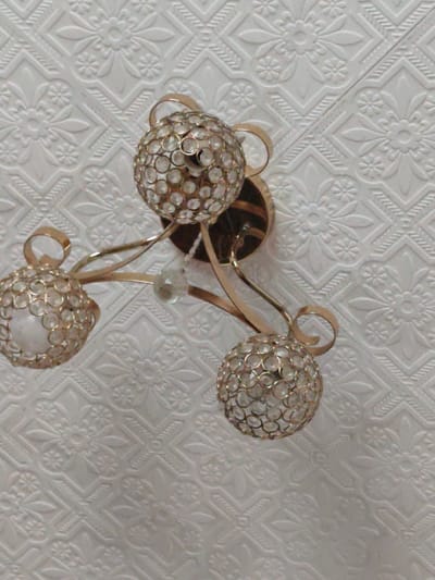 The image shows a close-up view of a decorative ceiling light fixture against a white, textured ceiling. The light fixture has a metallic gold finish and features three spherical lamps, each covered in clear, faceted crystals. These crystal-covered spheres are attached to curved metal arms that extend from a central mount. One of the spheres appears to be illuminated from within. A small, faceted clear prism dangles from one of the arms between two of the spheres. The ceiling has a repeating embossed pattern of floral and geometric shapes. The scene is static and appears to be indoors. There are no people or any other objects visible in the image. The location of the image within the city of Karaganda, Russia, cannot be determined from the visual information provided.