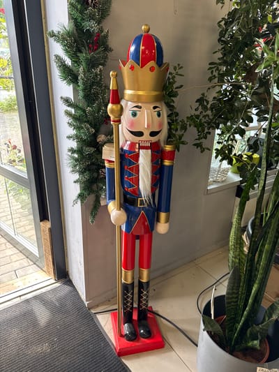 A tall, traditional nutcracker figure stands prominently in an indoor setting, likely a shop or entryway. The nutcracker is dressed in a blue jacket with red trim and gold accents, red trousers, and a crown. It holds a golden staff. To its left, a green garland adorned with pinecones and red berries is draped. Behind the nutcracker, a large leafy plant is visible, its branches cascading downwards. The floor is tiled, and a dark grey doormat is positioned in the foreground. The background includes a window with a view of greenery outside and a white radiator. The image appears to be taken at a slightly low angle.