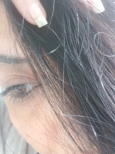 A close-up, high-detail forensic image captures the right side of a person's face, specifically their right eye and adjacent hair. The individual has dark, straight hair intermingled with prominent strands of grey/white hair. A manicured finger, featuring a square-tipped, light-colored nail, is observed lifting a section of this hair, exposing some of the grey strands. The visible right eye has a brown iris, dark pupil, and defined dark eyelashes. The eyebrow is also dark and discernible. The surrounding facial skin appears smooth. The immediate background is soft and brightly lit, offering no specific environmental identifiers. The location is in the city of Vernon, France.
