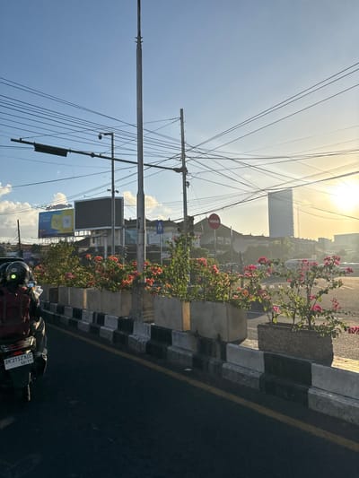 The image depicts a street scene in Denpasar Barat, Indonesia, during the golden hour. In the foreground, a motorcycle with a red backpack is parked on the left side of the road, facing away from the viewer. Its license plate reads "DK 2712 ACC 10-29". The road surface is asphalt, marked with a yellow line along the right edge. A raised planter with black and white striped concrete barriers runs along the right side of the road, filled with vibrant pink and orange flowering plants. In the midground, several buildings with traditional architecture are visible, along with a large billboard. The sky is a clear blue with some scattered clouds, and the setting sun casts a warm glow over the scene. Overhead, a complex network of power lines crisscrosses the sky. In the far background, a tall, modern building stands out against the horizon. The overall impression is one of a bustling urban environment bathed in the soft light of sunset.