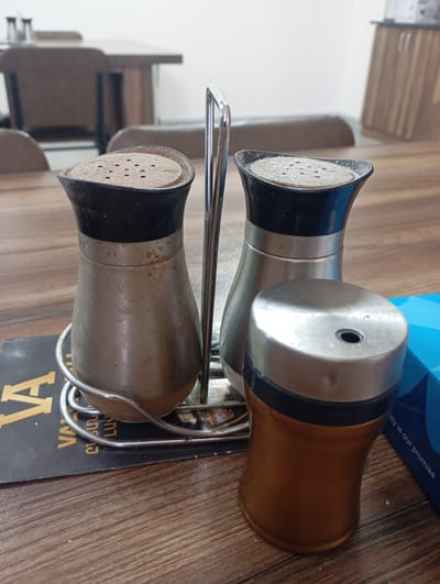 A salt and pepper shaker set, made of metal and black plastic, sits on a wooden table next to a blue box. The shakers have silver-toned bodies and black tops with holes for dispensing. The set is held together by a silver wire rack. A part of a black menu with yellow text is visible under the shakers. The scene appears to be in a restaurant or dining establishment, potentially in the city of Ararat, Armenia, given the style of furniture and overall ambiance. No people are present in the image.