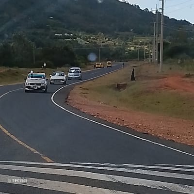 This image displays a two-lane asphalt road curving to the right through a rural area in Kola ward, Kenya. The road is marked with a solid white center line and a yellow line delineating the shoulder, with a partial zebra crossing visible in the immediate foreground.

Vehicular traffic is observed moving away from the camera. In the leftmost lane, a light-colored sedan with a blue object on its roof is followed closely by a white sedan, and then a light-colored minivan. Further down the road, on the right lane, three yellow vehicles, appearing to be vans or matatus, are in motion.

On the distant left shoulder, two individuals are visible, walking alongside the road. A solitary dark-clad figure stands on the right shoulder in the mid-ground, positioned near a low concrete structure or culvert.

The environment features reddish-brown soil shoulders and green grassy verges. The background consists of rolling green hills, sparsely covered with trees and scattered structures, extending under an overcast sky. A series of wooden utility poles with overhead lines run parallel to the road on the right side.