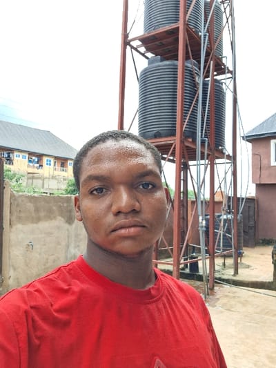 The image depicts an outdoor scene in the city of Ukwulu, Nigeria. A male subject, appearing to be of African descent, occupies the central foreground, facing the camera directly with a neutral expression. He has short, dark hair and wears a plain red short-sleeved t-shirt; his forehead and nose exhibit a slight sheen. The background features a tall, reddish-brown metal framework supporting three vertically stacked, black, ribbed plastic water tanks. Multiple grey PVC pipes are integrated into the structure, connecting to the tanks. The ground beneath the water tower is paved and wet. To the left of the subject, a light-colored concrete wall precedes a multi-story building with a dark corrugated roof, yellow-painted walls, and blue-framed windows and balconies. A brown building with a dark roof is partially visible behind the water tower on the right. The sky is uniformly overcast.