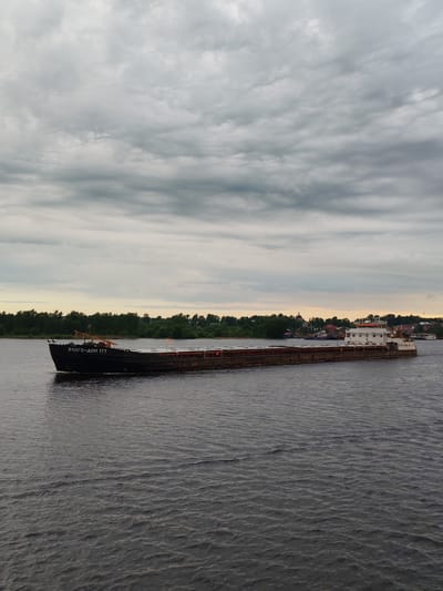 A long, dark cargo barge with the inscription "ВОЛГО-ДОН 177" on its bow, is moving across a dark body of water, likely the Volga River. On the barge's stern is a white, multi-tiered structure, likely the captain's cabin. The barge appears to be in the city of Myshkin, Russia, as suggested by the trees and buildings visible on the distant shore. The sky is overcast with grey clouds, creating a muted, somewhat gloomy atmosphere. Ripples are visible on the water's surface.