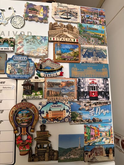 The image shows a collection of souvenir magnets affixed to a white refrigerator or other similar metal surface. The magnets are from various locations, mostly in Europe, including Granada, Salou, Istanbul, Nerja, Burano, Hita, Toledo, Asturias, Bulgaria, Venice, and other places in Italy and Spain. The magnets vary in shape, size, and style, ranging from simple rectangular magnets to more elaborate 3D designs. Some magnets feature landmarks, cityscapes, or country names, while others depict specific themes like food or local culture. A calendar with the word "Saturday" is partially visible at the bottom right of the image. The scene is well-lit and the magnets are arranged in a dense, overlapping pattern. The city of Azuqueca de Henares is not visible in the image.