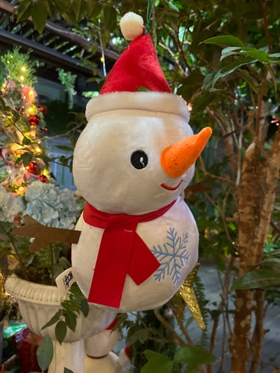 A plush snowman decoration is suspended by a green string amidst green foliage in Buôn Ma Thuột, Vietnam. The snowman, white in color, features a red Santa hat with a white pom-pom and brim, black embroidered eyes, an orange conical plush nose, and a red embroidered smile. A red plush scarf is tied around its neck, and a light blue snowflake design is embroidered on its lower body. Brown felt stick-like arms protrude from its sides. A product tag is visible on its left side. In the background to the left, a white fluted planter holds a plant adorned with numerous red spherical ornaments and warm yellow string lights, with light blue faux flowers visible below. To the right, green foliage and a tree trunk are present. String lights and a wooden pergola structure are visible in the deeper background. A partially obscured second snowman decoration and a golden star ornament are also visible.
