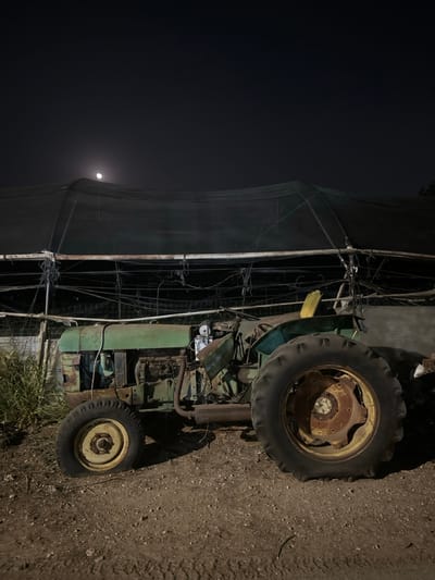 The image depicts a static scene at night in Kfar Monash, Israel, featuring a dilapidated green tractor positioned on a dirt ground surface. The tractor is severely weathered, exhibiting significant rust on its engine components, front axle, and wheel hubs, particularly on the front left wheel which is flat and appears deflated. Its body panels are faded green, and some are missing or damaged, exposing internal machinery. A yellow object, possibly part of a seat or a discarded item, is visible on the driver's side.

Behind the tractor, a large, dark mesh shade net structure, likely part of an agricultural facility or greenhouse, spans the background. This structure is supported by numerous metal poles and wires, many of which appear loose, tangled, or in disrepair. To the far left, a patch of green vegetation, possibly tall grass or weeds, grows from the ground. The sky above is dark, indicating nighttime, with a prominent, bright full moon visible in the upper-left quadrant of the frame, partially obscured by the shade net structure. No individuals are present, and no actions or interactions are depicted. The overall environment suggests a rural or agricultural setting, possibly abandoned or disused, under lunar illumination.