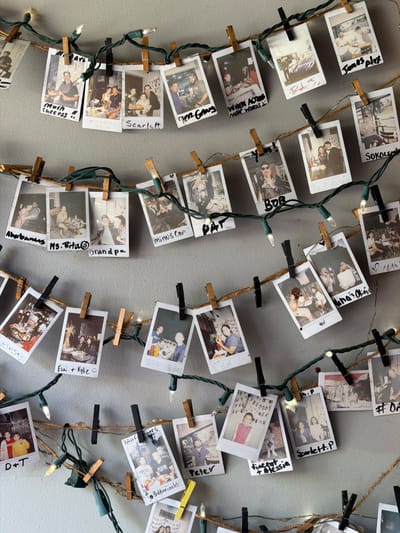 The image displays a wall decorated with a string of Christmas lights adorned with instant photographs. The photographs are clipped to the lights with wooden and black clothespins. The string of lights and photos is hung in a cascading manner, with multiple rows. The photographs feature various individuals in casual settings, some with handwritten captions. The wall appears to be painted a neutral gray color. The overall scene suggests a personal and decorative arrangement. There is no discernible indication of the city of Edgewater, United States of America.