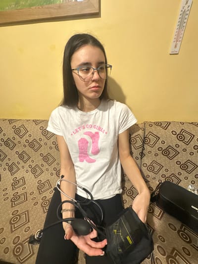An indoor scene in a residence located in Letnitsa, Bulgaria, depicts a young adult female seated on a patterned sofa against a plain light yellow wall. The female, with long dark hair and wearing light blue-framed glasses, is oriented towards the viewer but looking towards her left. Her mouth is slightly downturned. She wears a white t-shirt featuring a "LET'S GO GIRLS" text and a graphic of two pink cowboy boots, and dark pants.
In her right hand, she holds the black rubber bulb component of a sphygmomanometer. The associated black blood pressure cuff, displaying "SODAS" branding and yellow markings, rests on her lap/left leg. A stethoscope, with silver ear tubes and black tubing, is draped over her left forearm. A woven bracelet is visible on her left wrist.
To her right on the sofa, a black zippered case is present. The wall behind her features a portion of a framed picture with a wooden frame visible in the upper left and a white wall thermometer displaying both Celsius and Fahrenheit scales, labeled "MADE IN CHINA," positioned towards the upper right. The sofa has a repeating pattern of geometric shapes, including squares and circles, in shades of brown.