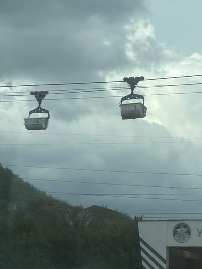 The image shows two cable cars hanging from cables against a cloudy sky. The cable cars are branded with the letter 'S' and the number '229'. In the background, there's a mountainside covered in greenery. Part of a sign or building with a circular emblem can be seen on the right. It's Kipra, Bulgaria.