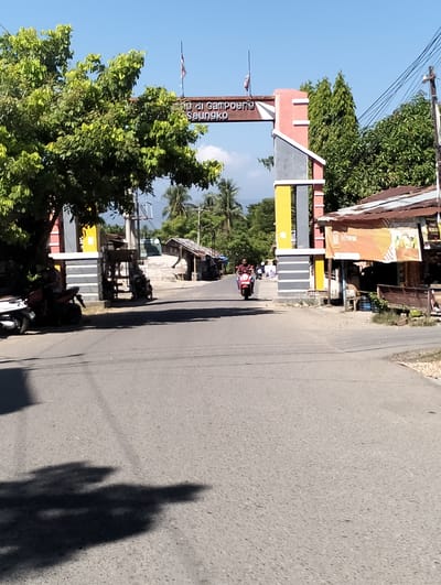 This image depicts an outdoor scene on a clear, sunny day, likely in Keudejunieb, Indonesia, suggested by the presence of an archway with Indonesian text. The archway, constructed with red, gray, and yellow elements, spans across a paved road. The text on the archway reads "Gampong Seunöngko," indicating the name of a village or settlement.
In the foreground, a portion of the road is visible, with shadows from trees cast upon it. To the left of the road, a motorcycle is parked near a large tree with lush green foliage. Further down the road, a person is riding a red scooter, approaching the archway.
To the right of the road, a building with a corrugated metal roof is visible, with an orange banner displaying the word "Miswar" in prominent lettering. Several smaller structures and trees are present in the background. Utility poles and overhead wires are also visible, indicating the presence of infrastructure. The overall environment appears to be a semi-rural or suburban area.