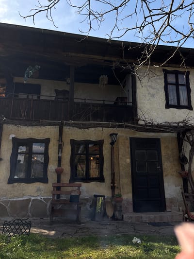 Exterior shot of a building in Gorno Trape, Bulgaria, featuring a three-story structure with a facade displaying textured plaster and dark wooden window frames. There are at least three visible windows, each with a distinct dark outline. A dark-colored front door is centrally located on the second level. A dried vine climbs the front of the building. The architecture includes a balcony or elevated walkway on the second story. Bare tree branches are visible in the upper left. A patch of grass and a stone-paved pathway can be seen to the right. Various decorative items are mounted on the walls and along the facade.