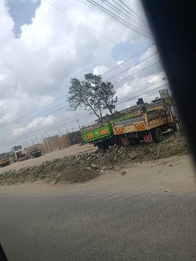 The image portrays an outdoor scene in Matungulu west ward, Kenya. A grey asphalt road occupies the foreground, leading to a dirt and rubble-strewn shoulder on the right. Two open-bed trucks are parked on this elevated, uneven ground. The left truck features a green cab and bed with yellow stripe accents; the right truck has a light brown cab and bed with yellow and red stripe accents. Both trucks display rear mudflaps. In the mid-ground, a green-leafed tree stands between the trucks and the background. Further behind, multiple other trucks are visible, along with piles of what appear to be bricks or construction materials, suggesting a building or storage site. Overhead power lines span the upper portion of the frame against a sky with significant cloud cover. No individuals are visible. The perspective is slightly tilted and appears to be from inside a vehicle due to a dark obstruction on the right.