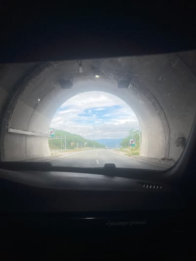 The image, taken from inside a vehicle looking out through the windshield, captures the exit of a tunnel onto an open road. The tunnel walls are made of concrete, with visible rebar and lighting fixtures. Outside the tunnel, a paved road stretches into the distance, flanked by green, tree-covered hills under a blue sky with scattered clouds. Several traffic signs are visible, including one with a green background and a white horizontal bar, indicating the end of a restriction. The foreground of the image shows the dashboard of the vehicle, with a textured surface and a small text label that reads "passenger princess." The overall scene suggests travel through a mountainous region. Without further information, the specific location in Blatino, Bulgaria cannot be determined from the image alone.