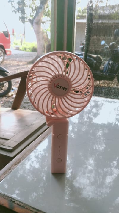 A close-up, eye-level shot shows a pink handheld fan with the brand name "VOTRE" on its front. The fan is placed on a white, glossy surface that reflects the environment. The background is slightly blurred, showing a red vehicle on the left and a person on a motorcycle on the right, suggesting an outdoor or semi-outdoor setting in Ketoyan, Indonesia. The overall lighting indicates it is daytime.