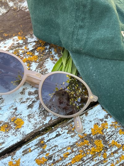 The image features a pair of light-colored sunglasses resting on a weathered wooden surface. The wood is painted a pale blue, with patches of peeling paint and textured yellow lichen, suggesting an outdoor setting. To the left of the sunglasses, a portion of a dark green cloth, possibly a hat or bag, is visible. Green foliage peeks out from beneath the cloth and the sunglasses. The lenses of the sunglasses reflect the surroundings, showing a clear sky and the branches of trees. On one of the sunglasses' arms, the text "P.Y.E" is visible in gold lettering. The lighting suggests it is daytime, with sunlight illuminating the scene and creating reflections. There is no discernible activity taking place other than the objects being placed on the surface. The overall impression is one of casual repose in an outdoor environment, possibly during warm weather. The location context of Zvonarevka, Russia, does not provide specific visual cues within the image itself.