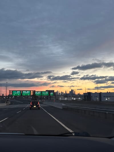 The image displays a multi-lane highway scene at dusk or dawn, viewed from the interior of a vehicle, with the dashboard visible at the bottom. A dark-colored SUV with illuminated taillights travels in the center lane ahead. Overhead gantries support four green highway signs with white text. From left to right, these signs indicate: "EXIT 15E Newark Airport Port Newark Elizabeth" (right arrow), "EXIT 15X Newark Downtown Holland Tunnel" (left arrow), "EXIT 15W Newark Airport" (right arrow), and another sign partially visible with text including "Jersey City Holland Tunnel." To the right of the highway, a barrier separates the road from industrial or commercial buildings. In the distance, an urban skyline is silhouetted against a sky characterized by orange, yellow, and grey cloud formations. The scene is located on the New Jersey Turnpike (I-95) in or immediately adjacent to the city of Newark, New Jersey, United States, indicating travel generally eastward towards Jersey City and the New York metropolitan area.