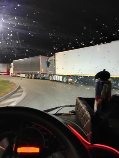 The image was taken from inside a truck's cab at night, with raindrops visible on the windshield. A line of semi-trailer trucks is parked or moving slowly on a road that curves to the left. The truck in the foreground has a dark dashboard with a lit orange digital display showing "699996 14:05". Next to the dashboard, a Red Bull can is visible. The road surface appears wet. In the background, the headlights of other vehicles and illuminated signs are visible, suggesting a rest stop or industrial area. The scene is consistent with the location of the D1 highway near Drahelčice, Czechia, a common place for truck stops and traffic congestion.