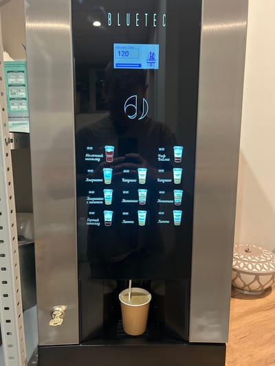 The image shows a BLUETEC beverage vending machine in what appears to be a public space. The machine has a digital display that shows "120" in large numbers, with the text "Капучино 180мл" above it. Below the display, there is a selection of drink options in Russian, each with an associated cup icon. Some of the drink names visible include "Молочный шоколад", "Американо", "Капучино", "Моккачино", and "Латте". A cup is currently being filled with a light-colored liquid from the dispenser. To the left of the machine, a white metal shelving unit is visible, with some printed material attached to it. The machine is situated on a wooden surface, and a decorative bowl is visible to the right. There is no specific information or visual cues in the image that indicate the location is Postol, Russia.