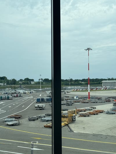 This image presents a daytime view of an airport tarmac at Malpensa Airport in Ferno, Italy, likely seen from an elevated position within a terminal building, framed by a dark horizontal bar in the foreground.
The scene is dominated by a wide array of ground support equipment (GSE) parked across the concrete and asphalt surfaces. Numerous baggage carts and cargo dollies, some empty and others appearing to hold cargo, are neatly arranged in rows. A yellow ground power unit (GPU) or pushback tractor is prominent on the right side. The tarmac is clearly marked with white and yellow lines, including directional arrows, indicating operational zones and movement paths.
In the mid-ground, a tall, slender pole, painted with alternating red and white stripes and topped with several sensors or antennas, stands out against the sky. Beyond the immediate tarmac, a lush line of green trees provides a natural boundary, with hints of distant buildings behind them. The sky is overcast with diffuse white clouds, suggesting a bright but not sunny day.
No people are visible in the frame, and while equipment is present, no active movement or specific event is taking place, implying the equipment is staged or parked between operations. There is no clearly legible text visible on any of the objects or ground markings.