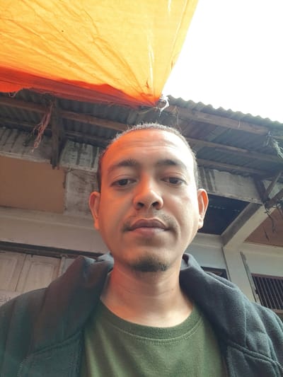 A man is photographed from a low angle, wearing a green t-shirt and a dark jacket. Above him, an orange sheet or tarp is visible. Behind the man, the roof of a building with corrugated metal is seen, along with weathered wood and a small section of a white structure with a barred window. The lighting suggests an outdoor setting on a possibly overcast day.