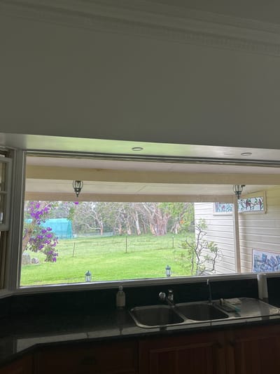 A kitchen sink occupies the right side of the frame, situated beside a window showcasing a vibrant green lawn. Beyond the lawn, a sparse woodland area is visible. The window is adorned with decorative exterior lights hanging from the eaves of a porch. The left side of the image consists of a light gray wall with intricate molding. The outside scene is partially obscured by the window frame and porch structure.