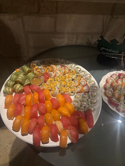 The image shows two platters of sushi on a dark gray surface. The larger, white platter contains a variety of sushi types: California rolls, spicy tuna rolls, cucumber rolls, and nigiri with both salmon and tuna. A small container of sauce sits amongst the sushi. The smaller platter also contains various sushi rolls. In the background, there's a "graduate" decoration and a brick wall. The scene appears to be indoors, possibly in a home or event space. This image does not have enough details to confirm its location in Southlake, United States of America.