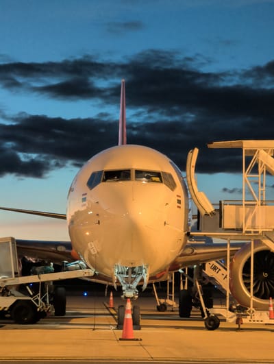 This media file shows the front of a large passenger airplane parked at an airport gate. The airplane is white with a pinkish-purple tail fin. A jet bridge is connected to the aircraft's door, indicating passengers are likely boarding or disembarking. The scene is viewed from a low angle, emphasizing the size of the plane.
The setting appears to be an airport tarmac at dusk or dawn, as evidenced by the twilight sky with dark, dramatic clouds. Artificial lighting from the airport infrastructure illuminates the aircraft and surrounding area, creating a warm glow. In the background, the large engine of the airplane is visible, along with parts of the jet bridge structure, including wheels and support beams. Orange traffic cones are placed near the aircraft's landing gear, a common safety measure at airports.
One visible piece of text on a white panel reads "-URT- P", likely part of a registration or identification marking for the aircraft or ground equipment. The overall mood is one of activity at an airport, with the airplane poised for its next journey. The presence of clouds suggests potentially changing weather conditions, but the immediate area appears clear. The time of day is implied by the transition from daylight to darkness, creating a visually striking contrast.
