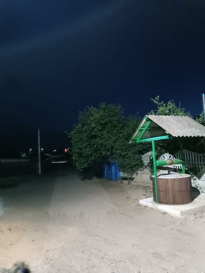 A well is pictured at night in a rural area, possibly in Leușeni, Moldova, given the context of the query. The sky is a deep, dark blue, indicating nighttime. In the foreground and to the right, a rustic well with a wooden barrel sits on a concrete base, covered by a sloped, grey, corrugated roof supported by green metal posts. A green mechanism for drawing water is visible. Behind the well, a white picket fence is partially obscured by lush green foliage. To the left, a dirt road stretches into the distance, lined with sparse vegetation and distant lights. A tall, light-colored pole stands near the road, and a blue structure is visible behind some trees. The overall scene is quiet and appears to be a residential or agricultural setting.