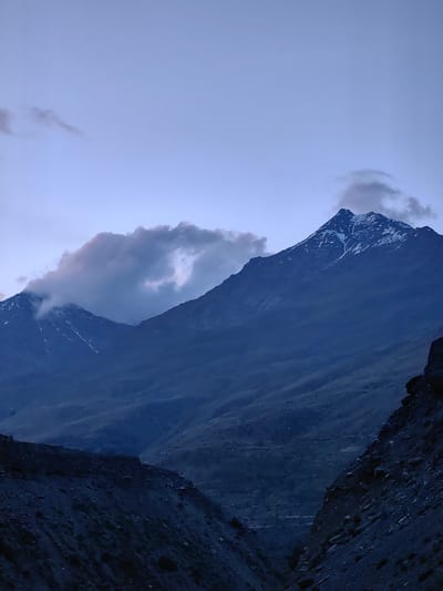 The image displays a majestic mountain range under a twilight sky. The peaks are dusted with snow, and dramatic clouds swirl around them, hinting at atmospheric conditions. The foreground is dominated by rocky, dark terrain, creating a sense of depth and ruggedness. The scene evokes a feeling of remote, natural beauty, typical of the high-altitude regions of India.