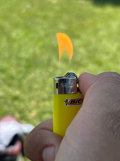 The image shows a close-up of a person's hand holding a yellow BIC lighter, which is lit, producing a small orange flame. The background is a blur of green, suggesting an outdoor setting with grass. The lighting appears bright, indicating it is daytime, possibly sunny. Visible text on the lighter includes "BIC" and a small graphic of a stick figure. The context of Vicenza, Italy, does not provide specific visual cues within the image itself, but it situates the photograph geographically. The focus is on the lighter and the flame, with the hand and background providing supporting context.