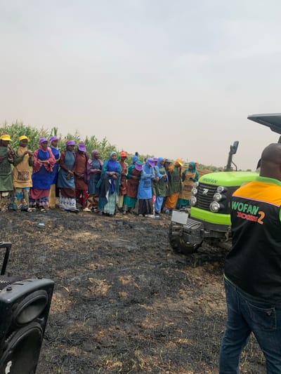 The image depicts a gathering of people in a field in Ringimawa, Nigeria, located at coordinates 11.666 latitude and 8.414 longitude. The field appears to be recently burned with black ash covering the ground, with a few green plants emerging. A large group of women, all wearing colorful clothing and head coverings, stand in a line facing a green tractor with a "WOFAN" logo. The tractor is parked on the edge of the field, facing the group of women. The women all appear to be interacting with one another, possibly engaged in a social event. A man, partially visible, is standing on the right edge of the image, with a black "WOFAN ICON2" shirt, wearing blue jeans. He is standing beside the green tractor and looking towards the group of women. A large speaker is in the foreground of the image, facing the women, suggesting that the gathering may involve music. The image appears to be taken during the daytime with cloudy skies.