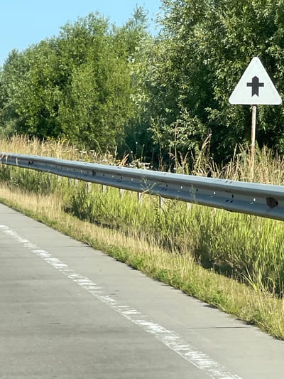 A white triangular road sign with a black symbol resembling an arrow pointing forward and splitting into two is visible on a metal pole. The sign is positioned near a metal guardrail, with a paved road and white lane markings in the foreground. Lush green trees and tall grass line the side of the road, suggesting a rural or semi-rural environment. The sky is clear and blue, indicating fair weather. The location is identified as Stremyhorod, Ukraine.