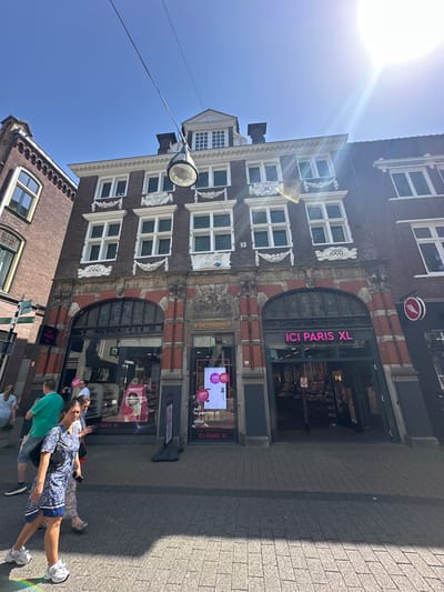 This media file depicts a sunny day in Zwolle, Netherlands, as suggested by the strong sunlight and clear blue sky. The scene is dominated by historic Dutch architecture, with a focus on a multi-story building featuring ornate details, multiple windows, and a prominent store with the signage "ICI PARIS XL". Several people are walking on the tiled sidewalk in front of the store, casting shadows that indicate the time of day is likely midday or early afternoon. The people appear to be casually strolling, some carrying bags, suggesting a normal day of shopping or sightseeing.

Visible text includes store names like "ICI PARIS XL" and "CHANEL," along with promotional text such as "TOP DEAL," "-35%," and "25% DEAL," indicating commercial activity. The building itself has inscriptions like "1600" and "ANNO," hinting at its historical significance. A street lamp hangs from a wire across the street, adding a functional element to the urban landscape. The overall impression is one of a vibrant, historic European city on a bright, clear day.