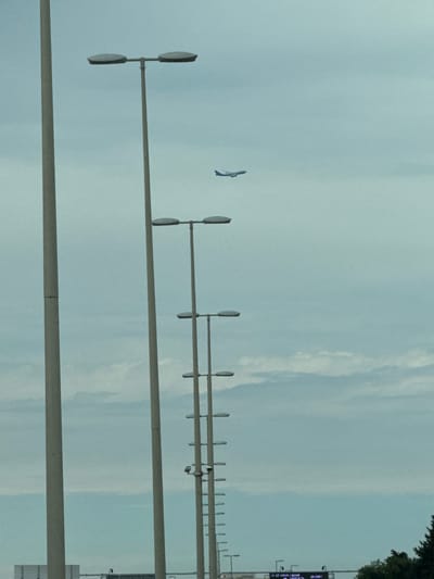 In Khazar Raion, Azerbaijan, an airplane is captured in flight against a cloudy sky. The image features a series of tall lampposts lined up in the foreground, extending vertically through the frame. The lampposts are equipped with horizontal lights, creating a recurring pattern. In the distance, the airplane is visible, a small silhouette against the overcast sky. The overall scene depicts an urban or suburban environment with infrastructure for lighting alongside the natural element of the sky and a passing aircraft.