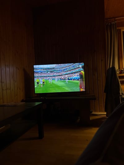 A television is on in a dimly lit room, displaying a soccer match. The walls are paneled with wood, and a curtain hangs on the right side of the frame. There is a dark wooden table in the foreground, and a jacket is hanging on the wall behind the television. The floor is made of wood. The image does not provide any information to determine the location in the city of Bykovo, Russia.