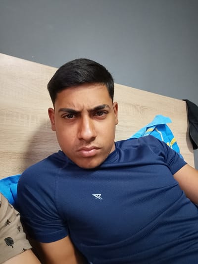 The image displays a young adult male, located indoors in Zagore, Bulgaria. He is positioned facing forward, looking directly at the camera with a serious expression, characterized by furrowed eyebrows and slightly pursed lips. The subject has dark, short hair and a light stubble on his chin and upper lip. He is wearing a dark blue, short-sleeved athletic shirt with a subtle textured pattern and a white, stylized logo on the left chest. Behind the subject's left side is a light brown, wood-grained surface, consistent with a headboard or wall panel. Above this wooden surface and extending across the upper background is a plain, medium grey wall. To the subject's right, a crumpled blue towel with yellow striping is visible behind his shoulder, and further to the right, a dark-colored garment is draped or hanging. A small portion of a light-colored fabric with dark, leaf-like patterns is present in the lower left foreground.