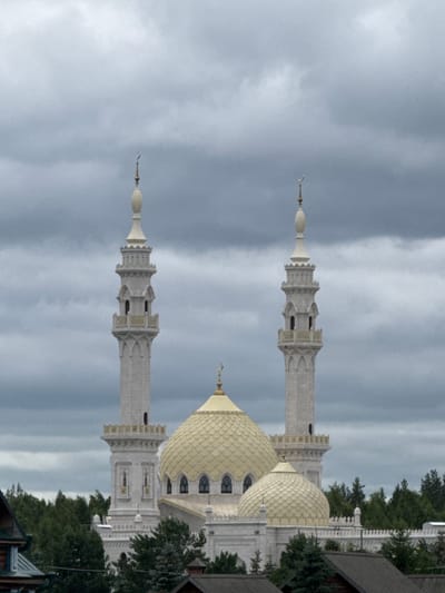 The image shows the White Mosque in Bolgar, Tatarstan, Russia, under a cloudy sky. The mosque is a large, white stone structure with two prominent minarets flanking a central dome. The minarets are tall and slender, with decorative balconies and pointed finials topped with crescent moons. The main dome is golden and ribbed, and there are smaller domes on the roof. The mosque is surrounded by lush green trees, and in the foreground, parts of other buildings with dark roofs are visible. The overall atmosphere is serene and solemn.