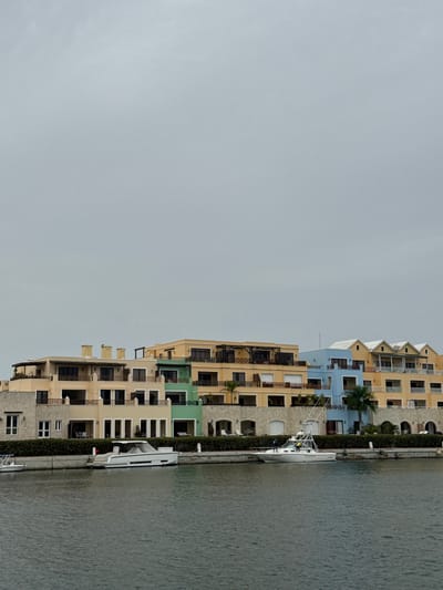 This image depicts a waterfront scene in Higüey, Dominican Republic, featuring a line of multi-story buildings situated along a canal. The buildings are architecturally diverse, with sections colored in shades of yellow, light blue, and green, adorned with balconies and numerous windows. A white boat with a prominent mast is docked alongside a hedge lining the edge of the canal, in front of one of the buildings. The water appears calm, reflecting the overcast sky. The scene suggests a residential or resort area. The overall mood is serene, with no visible activity or people. The weather appears cloudy. No text is visible in the image.