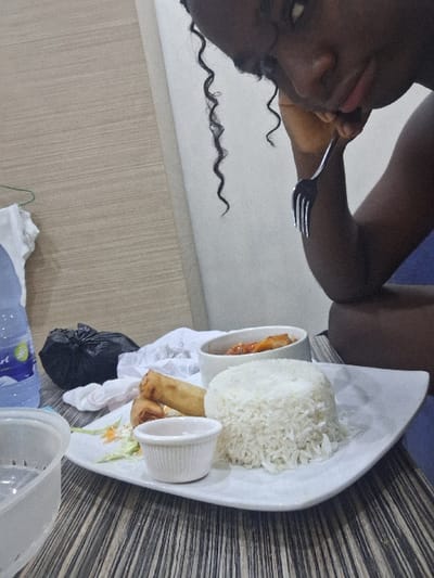 The scene is set indoors, depicting a meal taking place in the city of Close, Nigeria. A dark-skinned individual, partially visible from the chest up on the right, with dark, curly hair, holds a black-handled fork in their right hand, resting their chin on their left hand. The primary subject is a meal served on a striped wooden table. A white, rectangular plate in the foreground holds a conical mound of white rice, two golden-brown spring rolls with a small garnish of shredded green and orange vegetables, and a small white ramekin containing a light-colored dipping sauce. To the left of the main plate, a clear plastic bottle containing water with a blue label (possibly "Everest") is visible. Behind the water bottle, a crumpled black plastic bag and a piece of crumpled white fabric are present. A separate white bowl behind the main plate contains an orange-red dish with visible pieces of protein. A clear plastic container is positioned in the lower left corner. The background features light-colored walls; the left wall exhibits a subtle horizontal wood grain pattern, while the right wall appears plain.