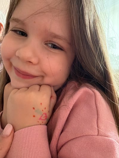 In Valozhyn, Belarus, a young girl with long, light brown hair is captured in a close-up, portrait-style photograph. She is leaning on her hands, which are positioned at her chin, and she is smiling gently, with her eyes looking slightly off-camera. Her left hand has a drawing on it, appearing to be a pink heart with some red dots and a light blue shape surrounding it. She is wearing a pink sweatshirt with ribbed cuffs. The lighting suggests an indoor setting, possibly near a window due to the soft, natural light illuminating her face and hair. The background is out of focus, with hints of a pale, possibly white or light-colored wall or curtain.