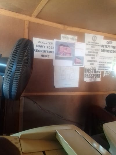 The image shows the interior of a small office space, likely a shop or a business, located at the coordinates 7.062 latitude, 6.273 longitude in Auchi, Nigeria. The wall is covered in a brown plywood panel, adorned with several posters and notices. A black electric fan with a metal casing is attached to the wall, its blades are still, suggesting it is not currently in use. The poster on the left reads "Register Navy 2023 Recruit me Here", while the center poster depicts a small two-story building. The poster on the right reads "Edo State Primary Healthcare Development Agency, Recruitment, Register for Federal Road Sa Recruitment 2023, Here!" It is further accompanied by two smaller posters with the text "Instant Passport Here" and "Take instant passport here". Below the posters, a large, rectangular, grey and white machine, possibly a copier or a printer, sits on a desk or a counter. The machine is partially obscured by the bottom edge of the image. The lighting in the room is dim. A section of another machine can be seen to the right of the image. The overall impression is of a functional space, likely serving a purpose related to the recruitment notices. The presence of the copier suggests this might be an office offering services related to paperwork and documentation. The image lacks any human figures, suggesting an empty shop or a moment captured when the staff were absent.