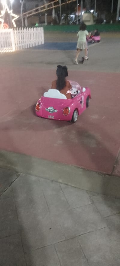 A young girl with dark hair sits in a pink toy car on a red paved surface. The car faces away from the camera. Another child on a scooter is visible in the background, moving to the right. In the distance, there is a lighted white picket fence and some trees. The bottom of the frame shows a gray, tiled surface. It appears to be an outdoor evening scene.