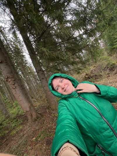 The image presents a woman in a green hooded winter coat standing in a forest. The background reveals tall, slender pine trees stretching upwards, with a view of the sky visible through the branches. The ground is covered with fallen pine needles and dry vegetation. The location is Protasy, Russia.