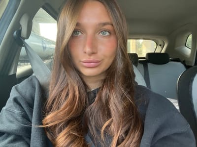 A young adult female is depicted in a selfie taken from the front passenger seat of a vehicle. She has long, wavy brown hair with lighter tones, light-colored eyes, and a light complexion featuring minor redness on her nose and cheeks. Her lips are slightly pursed. She is wearing a dark, long-sleeved sweatshirt and is secured by a grey shoulder seatbelt. The vehicle's interior shows light grey upholstery on the rear seats with dark headrests. Through the driver's side window, a large, white cylindrical tank with blue lettering and a logo is partially visible, suggesting an industrial or transport context. Through the rear passenger window, a blurred outdoor scene featuring trees and indistinct structures is visible. The specific location within La Fare-les-Oliviers, France, cannot be determined from the visual information presented.